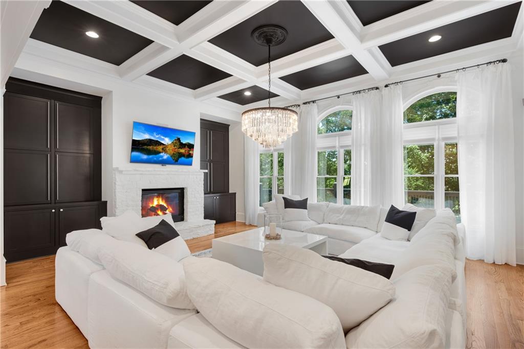 Great room of 3075 Wills Mill: a coffered ceiling painted dark with white beams, crystal chandelier, white sectional sofas around a stone fireplace, tall arched windows with sheer drapes