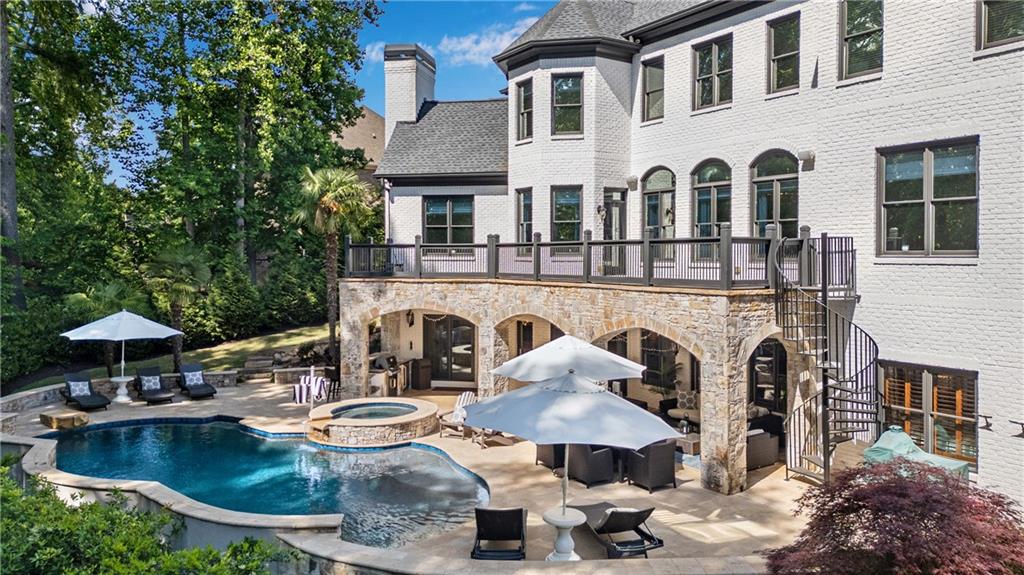 Rear elevation of 3075 Wills Mill in daytime: a three-level white-brick home with a turret bay, stone-arched terrace level, deck with railings, the saltwater pool and spa with umbrellas in the foreground, mature wooded lot behind