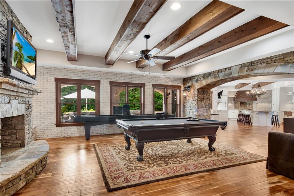 Terrace level recreation room at 3075 Wills Mill: full-size pool table on a Persian rug, shuffleboard table behind, rough-hewn wood ceiling beams, white-brick walls, stone-arched openings leading to the second kitchen, large windows looking out to the pool