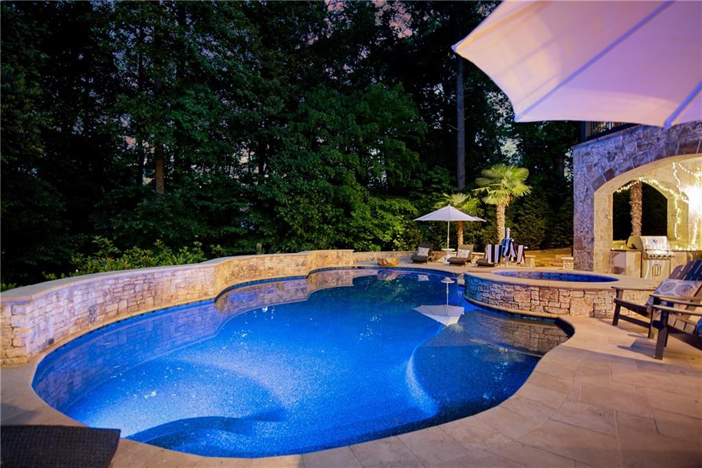 Saltwater pool of 3075 Wills Mill at dusk: freeform pool lit blue from within with a circular spa attached, stacked stone surround, travertine deck, mature pines and a half-open white umbrella to the side, palm at the far edge