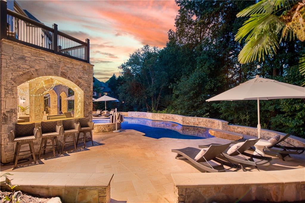 Twilight scene at 3075 Wills Mill: outdoor stone bar with wicker barstools under string-lit arches, white umbrellas over a chaise lounge cluster, the freeform pool reflecting the pink-orange sky, a Georgia sunset over the wooded backyard