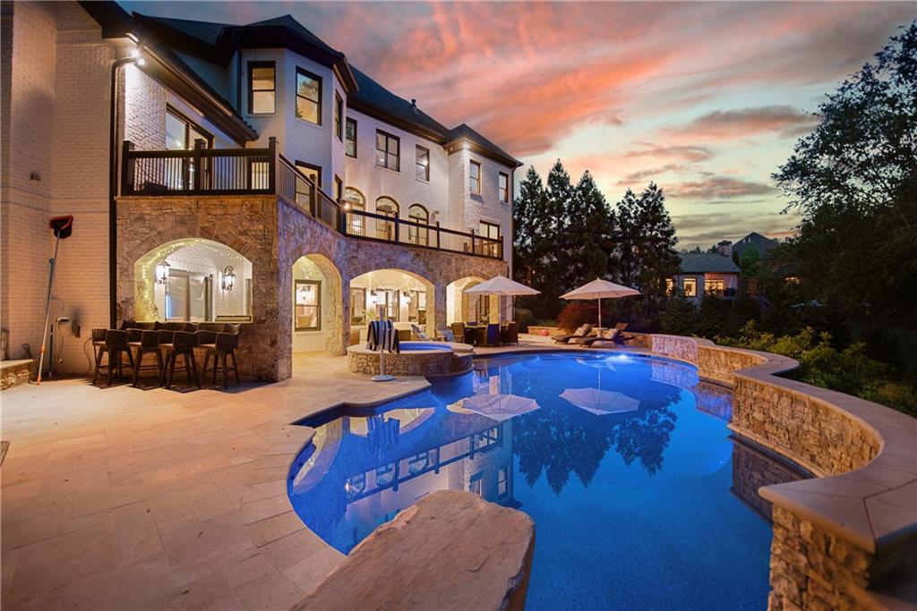 Rear elevation of 3075 Wills Mill Road at twilight: a saltwater pool lit from within, stone-arched terrace level under string lights, multi-level white brick estate with a pink-orange Georgia sunset sky
